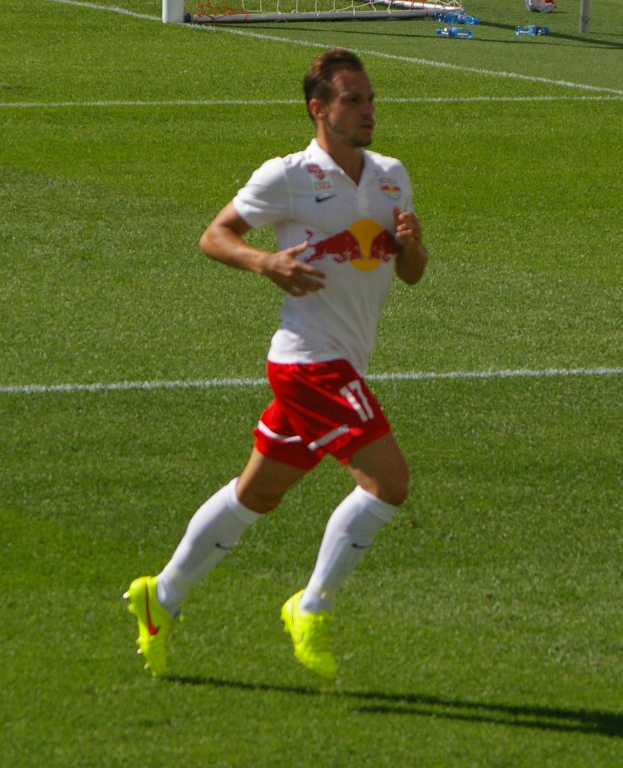 Ein Mann in weißem Hemd und roter Hose läuft auf einem Fußballfeld, im Hintergrund ist ein Tor zu sehen und ein paar Menschen stehen daneben, die Fußballschuhe tragen.