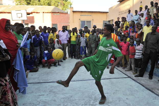 Ein junger Junge kickt einen Fussball vor einer Menge mit stehenden und sitzenden Menschen, mit Gebäuden, Bäumen und einem klaren blauen Himmel im Hintergrund.