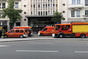 Eine Gruppe von Feuerwehrautos auf einer Straße in Paris geparkt, mit Menschen auf dem näheren Gehweg, Gebäuden, Bäumen und einem Fahrrad im Hintergrund.