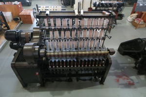 Eine Textilmaschine mit einem großen rechteckigen Rahmen und verschiedenen Knöpfen und Bedienelementen steht in der Mitte eines Raumes, umgeben von anderen Maschinen und Gegenständen, mit einer Tafel mit Text auf der rechten Seite und weiteren Maschinen im Hintergrund.