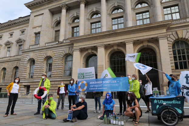Gruppe von maskierten Menschen mit Schildern vor einem Gebäude während einer Klimademonstration in Berlin, mit einem Fahrzeug und einem Geländer im Hintergrund.