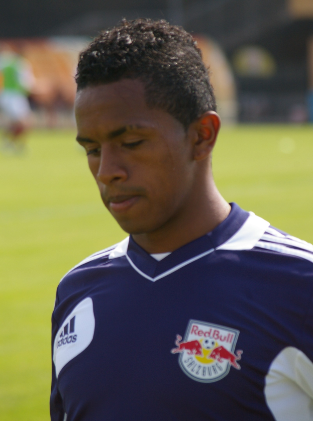 Junger Mann in blauem T-Shirt auf einem Fußballfeld mit entschlossener Miene, unscharfer Hintergrund.