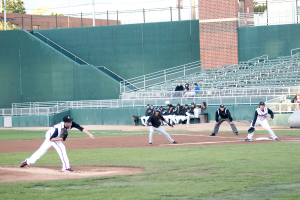 Baseball players are playing a game on a field, with spectators sitting on benches and chairs behind a railing, cars, a wall, pillars, and trees in the background.