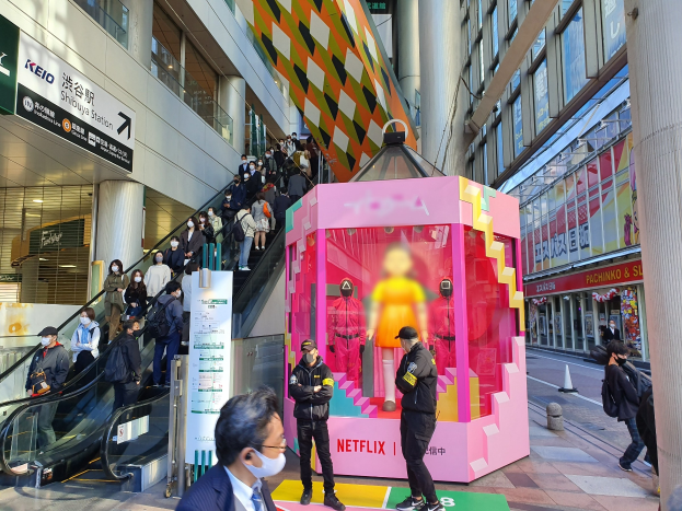 Eine Gruppe von Menschen steht vor einem Gebäude mit Glaswänden und Texttafeln, einige tragen Masken, während eine Rolltreppe mit Fahrgästen zu sehen ist; ein pinker Kasten in der Mitte enthält Schaufensterpuppen und trägt den Text "Netflix's neuer Netflix-Stand in Tokio."