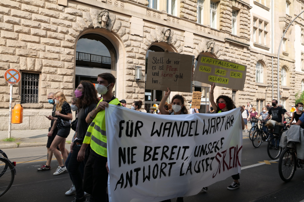 Eine Gruppe von Menschen, die während einer Demonstration in Berlin die Straße entlanggehen, einige halten Schilder und andere fahren Fahrräder, im Hintergrund ein Gebäude mit Fenstern, Bögen, Säulen und Skulpturen.
