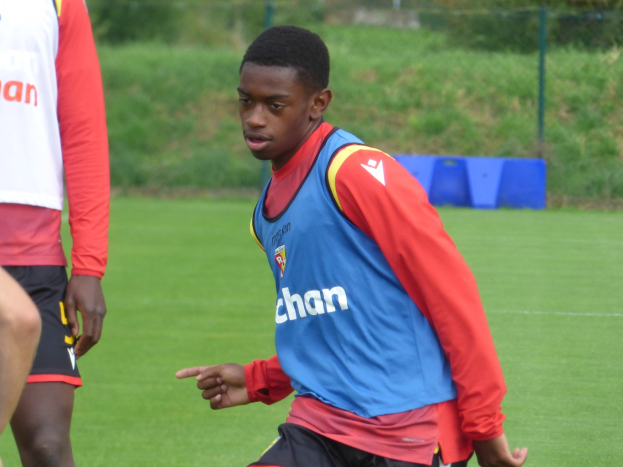Ein junger Mann in einem blauen Hemd und schwarzen Shorts, der auf einem Rasenfeld mit einem Zaun, Pflanzen und Bäumen umgeben Fußball spielt.
