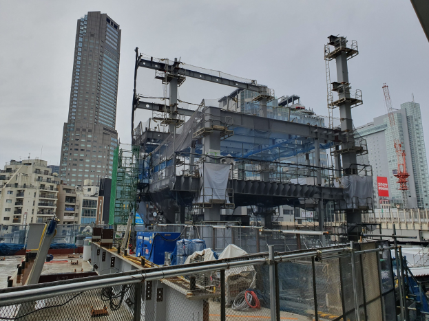 Großbaustelle für ein neues Kraftwerk in einer Stadt, umgeben von einem Metallzaun, mit umliegenden Gebäuden, Fahrzeugen, Polen, Drähten und einem bewölkten Himmel.