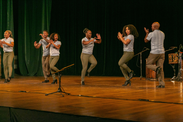Eine Gruppe von Menschen auf einer Bühne, die klatschen und feiern, mit Mikrofonen, Musikinstrumenten und einem Vorhang im Hintergrund.