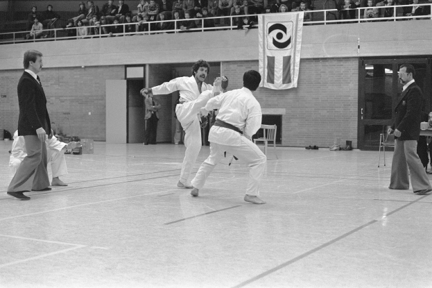 Schwarzes und wei├čes Bild einer gemischten Karate-Klasse in einer Turnhalle, mit einigen Teilnehmern, die stehen, und anderen, die sitzen, und einem Banner im Hintergrund.