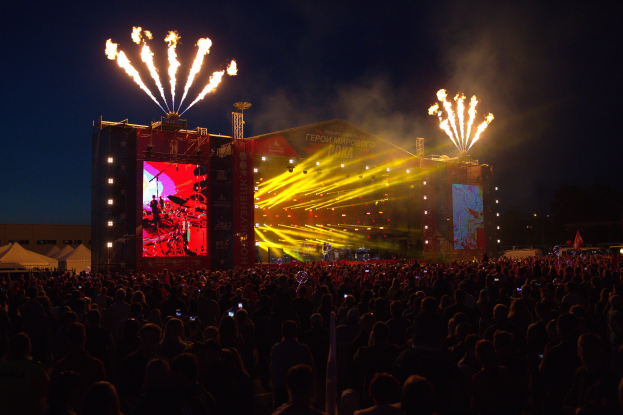 Großes Publikum bei einem Musikfestival mit einer Bühne, auf der Künstler unter hellen Scheinwerfern spielen, umgeben von Bannern und Zelten links und Feuerwerk am dunklen Himmel.