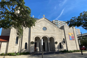 Die griechisch-orthodoxe Kirche im Osten von San Antonio, Texas, ist ein großer Bau mit Fenstern, Säulen, Bogengängen und Treppen mit Geländern, umgeben von Pflanzen, Bäumen und Fahrzeugen auf der Straße, unter einem bewölktem Himmel.