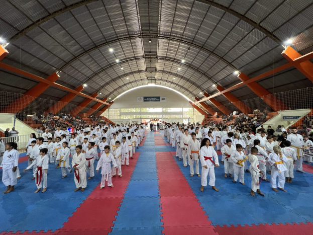Große Gruppe von Menschen auf einer blauen und roten Matte in einer Turnhalle mit Zuschauern auf Treppen und Deckenleuchten, Hintergrundplakate deuten auf ein Taekwondo-Event hin.