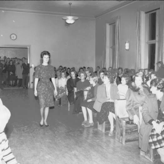Eine Frau in einem schwarzen und weißen Kleid läuft über einen Laufsteg während einer Modenschau für Frauenwahlrecht, mit einer Menge, die auf Stühlen sitzt und zuschaut, einer Uhr und Lampen an der umliegenden Wand und einer beleuchteten Decke.