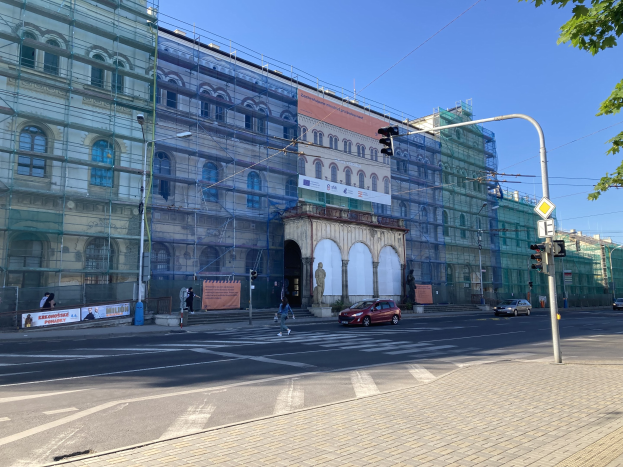 Großes Gebäude mit Gerüst während der Renovierung umgeben von Fahrzeugen, Fußgängern, Ampeln, Straßenschildern, Polen, Bäumen und einem klaren blauen Himmel.