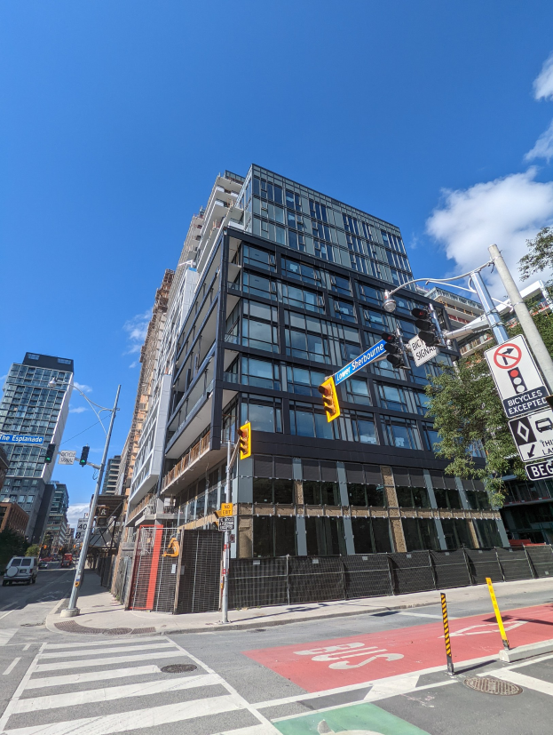 Eine belebte Stadtstraße mit einem hohen Apartment-/Reihenhausgebäude im Hintergrund, das zum Verkauf steht (541 East 57th Street, Unit 541), mit Fahrzeugen, Verkehrsampeln, Schildern, Bäumen, einem Zaun und einem bewölkten Himmel.