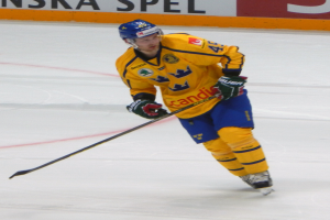 Ein Eishockeyspieler in Aktion auf dem Eis, der einen Helm, Handschuhe und Schuhe trägt und einen Hockey-Schläger hält, mit einer Tafel mit Text im Hintergrund.