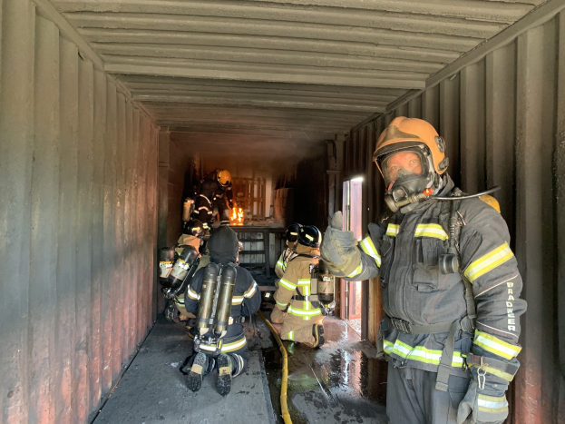 Feuerwehrleute in Schutzausrüstung in einem Container, einer hält ein Rohr, mit einem Feuerlöscher im Hintergrund.