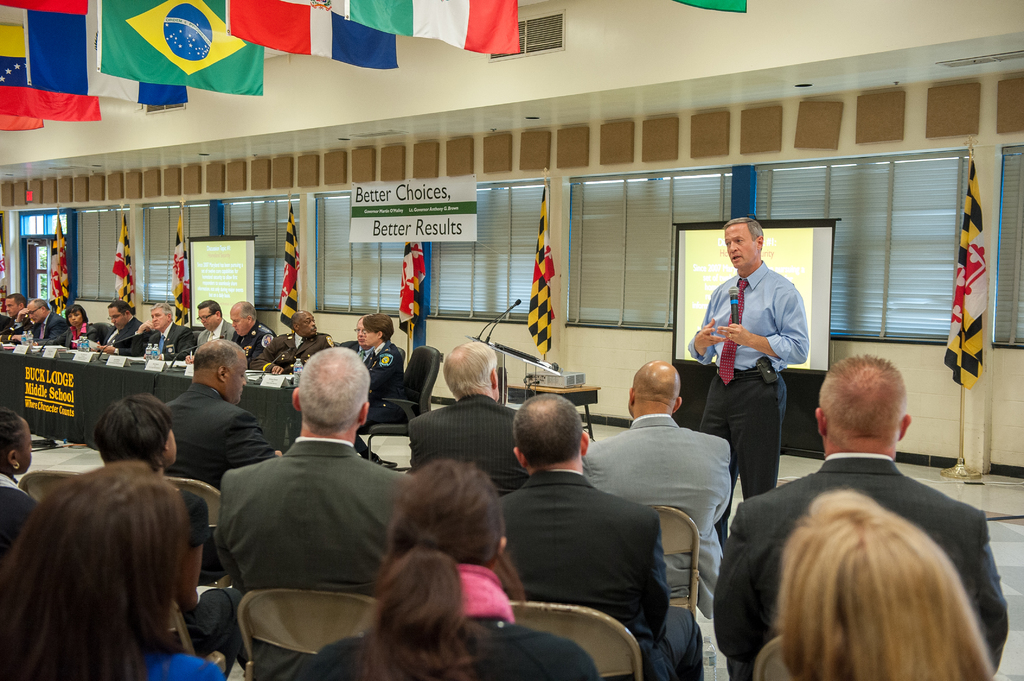 Eine Gruppe von Menschen, die auf Stühlen sitzen und einem Redner mit Mikrofon zuhören, mit einem Projektionsschirm hinter ihm, einem Projektor auf einem Tisch, einem Rednerpult mit Mikrofonen, einer Gruppe von Menschen mit Wasserflaschen, Fahnenmasten, hängenden Fahnen, verdeckten Fenstern und einer Tür auf der linken Seite.