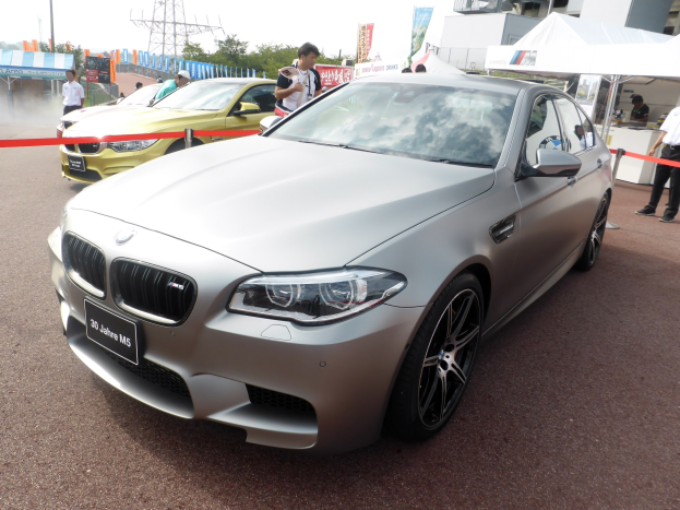 BMW M5 F10 M Performance Auto auf einer Straße mit Menschen, Zelten, Bannern, Bäumen, einem Turm und einem klaren blauen Himmel.
