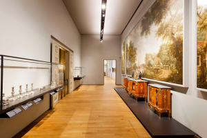 Ein Raum im Stedelijk Museum in Amsterdam, der verschiedene Kunstwerke, darunter Gemälde, Objekte, Texttafeln und eine Glasbox, mit Deckenbeleuchtung zeigt.