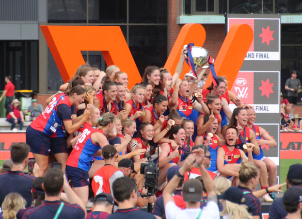Eine Gruppe von Frauenfußballerinnen in blauen und roten Uniformen feiert mit einem Pokal vor einer jubelnden Menge auf einem Rasenfeld, mit Zuschauern und beschrifteten Plakaten im Hintergrund.