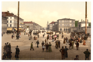 Ein altes Foto einer belebten Münchner Stadtstraße aus den frühen 1900er Jahren, das Fußgänger mit Schirmen, einen Pferdewagen im Vordergrund und Gebäude, Bäume und Pfähle im Hintergrund zeigt, mit Text am unteren Rand des Bildes.