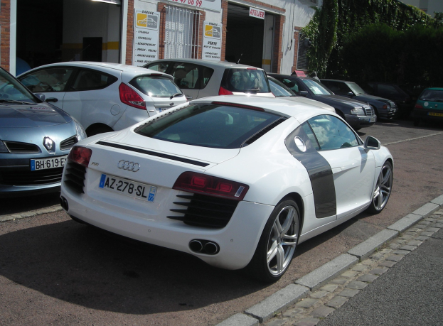 Ein weißer Audi R8 parkt vor einem Gebäude mit vernagelten Fenstern, umgeben von Bäumen auf der rechten Seite.