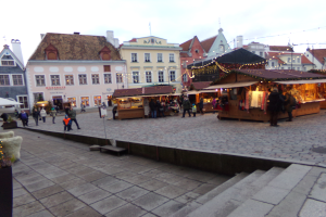 Ein lebendiger Weihnachtsmarkt in Tallinn, Estland, mit Menschen um dekorierte Stände, festliche Lichter und Gebäude mit Fenstern im Hintergrund, unter einem bewölkten Himmel, mit Stufen, die zum Markt führen, und Töpfen mit Pflanzen, die herumstehen.