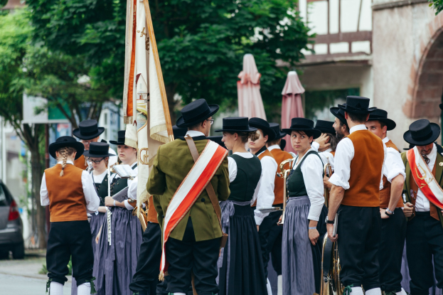 Eine Gruppe von Menschen in traditioneller bayerischer Tracht marschiert die Straße entlang, einige halten Musikinstrumente und Fahnen, im Hintergrund sind Bäume, Gebäude und ein Auto zu sehen.