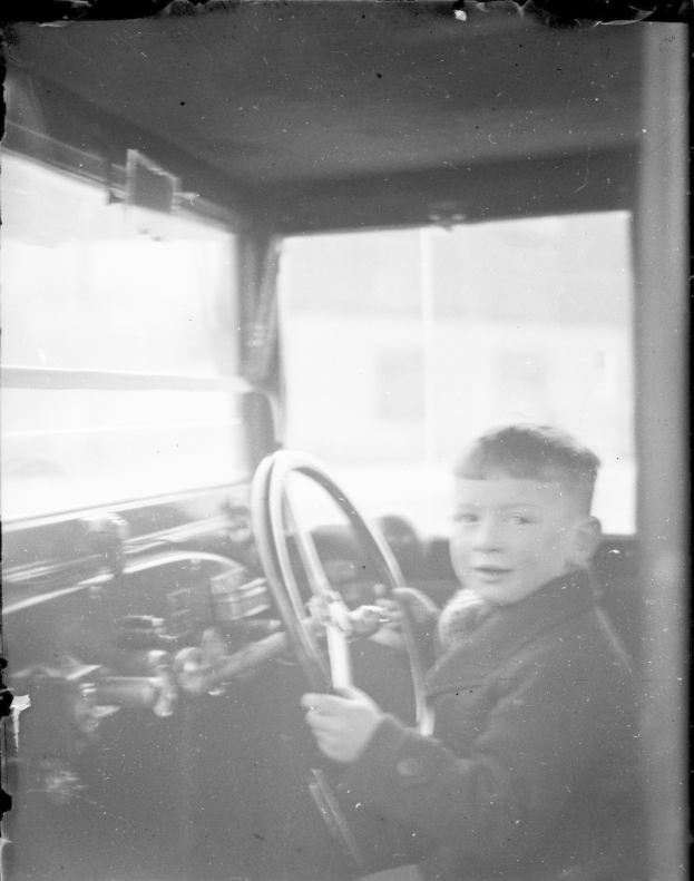 Ein junger Junge in Schwarz-Weiß sitzt im Fahrersitz eines Autos und hält das Lenkrad mit konzentrierter Miene fest.
