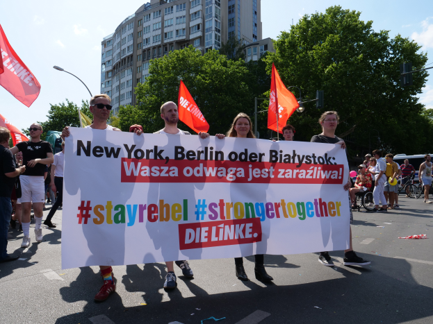 Eine Gruppe von Menschen geht eine von Bäumen gesäumte Straße in Berlin, Deutschland, entlang und hält ein Banner hoch, auf dem 'Stay Rebel Stronger Together' steht, mit Gebäuden und Laternenpfählen im Hintergrund und einem bewölkten Himmel.