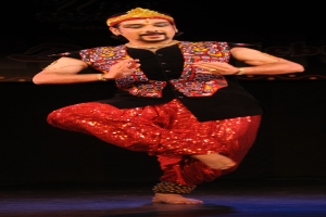 Ein Mann in roter und schwarzer Kleidung tanzt auf einer Bühne mit dunklem Hintergrund, seine Augen sind geschlossen und sein Mund leicht geöffnet.