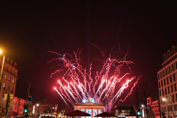 Eine belebte Stadtstraße an Silvester in Berlin, voller Menschen, Fahrzeuge und festlicher Dekoration, mit Feuerwerk, das den Himmel über den Gebäuden erhellt.