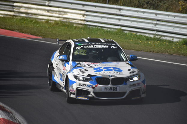 Ein BMW M4 DTM Rennauto auf einer Rennstrecke, umgeben von Gras, einem Zaun und Pflanzen, mit sichtbarem Text, Zahlen und einem Rad.