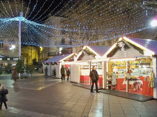Ein belebter Weihnachtsmarkt bei Nacht in einer Stadt, mit Menschen, die stehen, sitzen und ein Kind halten, geschmückt mit Lichtern, Pflanzen und Gebäuden unter einem bewölkten Himmel.