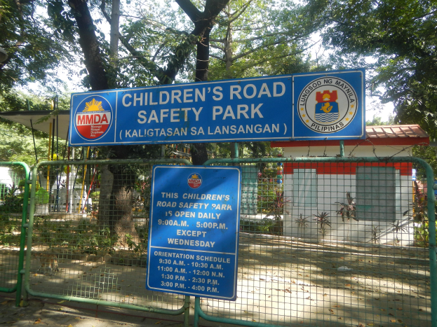 Metallzaun um einen Kindersicherheitspark mit zwei blauen Informationsschildern, einem kleinen Haus, Pflanzen, Bäumen und einem bewölkten Himmel.