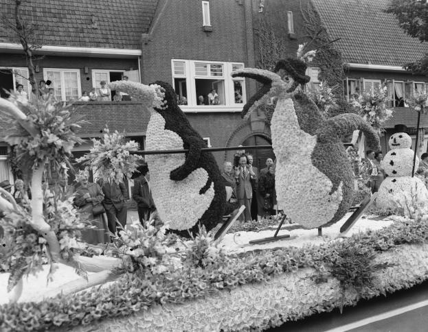 Schwarz-weiß-Foto eines Paradewagens mit Blumen und Pinguinfiguren, mit Menschen, Bäumen und Gebäuden im Hintergrund.