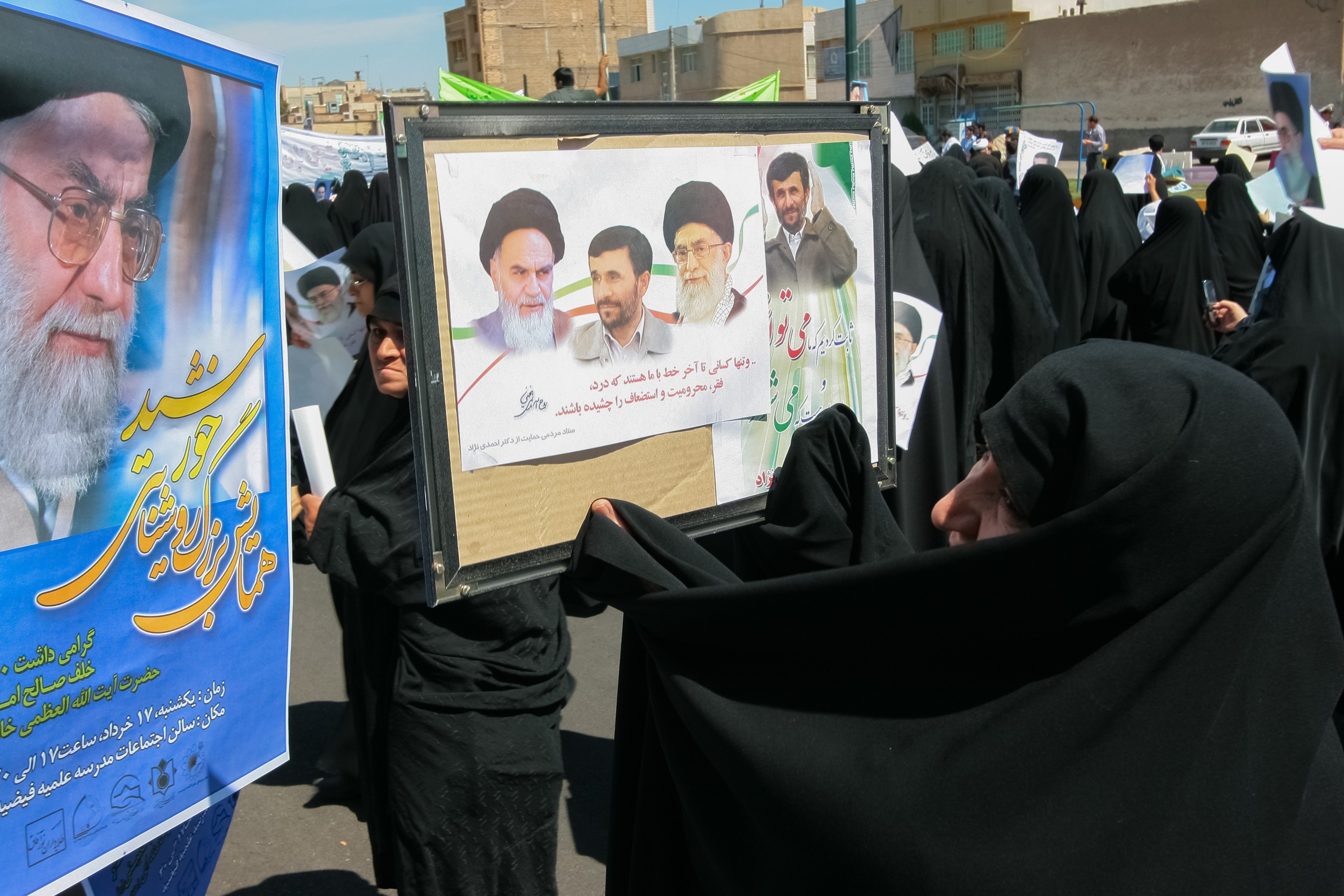 Eine Gruppe von Frauen in schwarzen Abayas hält Plakate mit Fotos von Ayatollah Ali Khamenei hoch, im Hintergrund sind Gebäude, Pfosten und Fahrzeuge unter einem klaren blauen Himmel zu sehen.