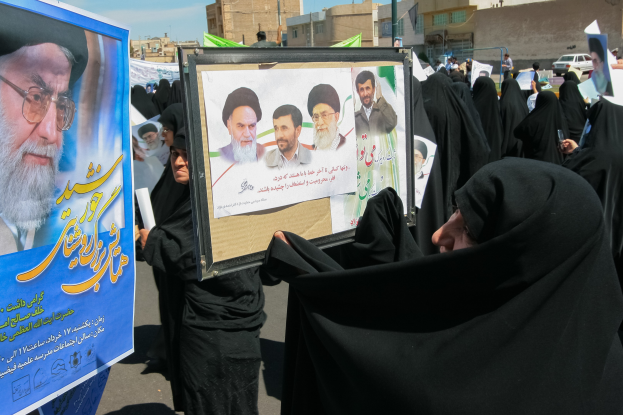 Eine Gruppe von Frauen in schwarzen Abayas hält Plakate mit Fotos von Ayatollah Ali Khamenei hoch, im Hintergrund sind Gebäude, Pfosten und Fahrzeuge unter einem klaren blauen Himmel zu sehen.