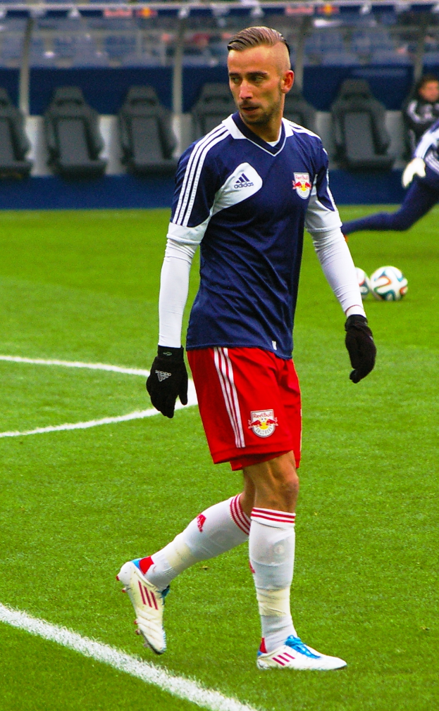 Ein Mann in einem blauen Hemd und roten Shorts spielt Fußball auf einem Feld, umgeben von Menschen, Stühlen und Pfählen.