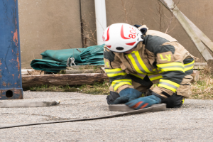 Ein Feuerwehrmann in Schutzausrüstung kniet auf dem Boden und verwendet einen Schlauch, um ein Feuer zu löschen, mit verschiedenen Gegenständen und einem Metallgegenstand im Hintergrund.