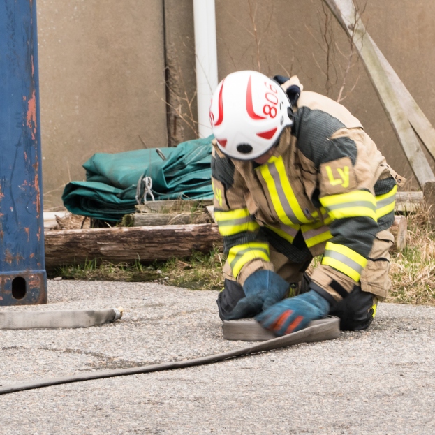 Ein Feuerwehrmann in Schutzausrüstung kniet auf dem Boden und verwendet einen Schlauch, um ein Feuer zu löschen, mit verschiedenen Gegenständen und einem Metallgegenstand im Hintergrund.