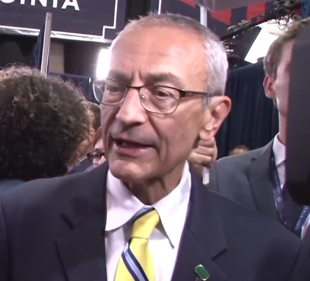 Ein Mann im Anzug und Krawatte, der eine Brille trägt, spricht zu Reportern mit einem Mikrofonständer vor ihm, umgeben von einer Gruppe von Menschen, mit Lichtern und einer Tafel im Hintergrund.