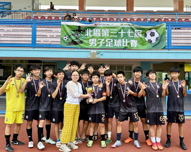 Gruppe junger Männer in Fußballtrikots auf einem Feld, mit Medaillen und einem Pokal in den Händen, mit einem Banner im Hintergrund, das 'Yokohama U-16 Boys Soccer Team' bezeichnet, und Zuschauern in der Nähe.