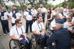 Eine Gruppe von Menschen im Rollstuhl, die vor einer Kulisse aus Bäumen, Pfählen, Gebäuden und einem klaren blauen Himmel diskutieren, wobei einige Mützen und Brillen tragen, was darauf hindeutet, dass sie an den Paralympischen Spielen teilnehmen.
