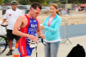 Ein Mann in einem roten und blauen Triathlonanzug läuft eine Straße entlang, neben ihm geht eine Frau, während im Hintergrund Menschen, Fahrräder, Verkehrskegel, Geländer, Fahrzeuge, Bäume und andere Objekte zu sehen sind.