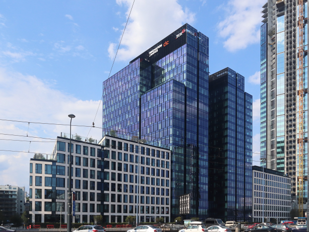Ein großes Gebäude mit vielen Fenstern, identifiziert als die Deutsche Bank-Zentrale in Frankfurt, Deutschland, umgeben von Straßeninfrastruktur, Fahrzeugen, Grünflächen und einem bewölkten Himmel.