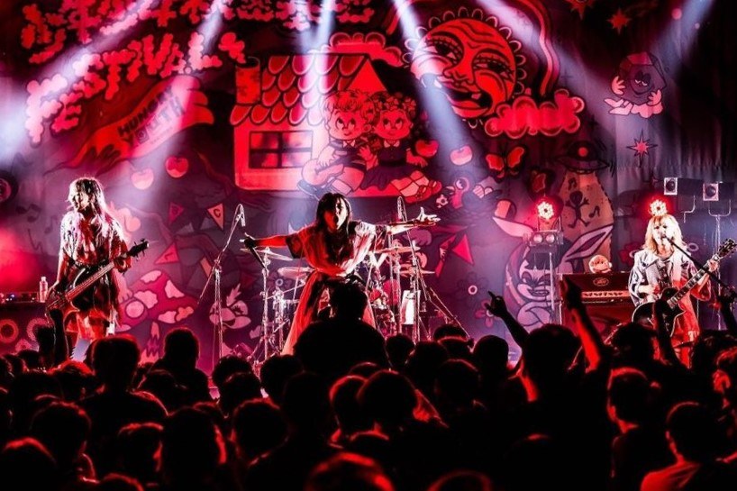 Ein Live-Auftritt der japanischen Rockband Korn auf der Bühne mit Musikern, die Gitarren spielen und ins Mikro singen, ein beleuchtetes Gemälde an der Wand dahinter und eine sichtbare Menge unten.