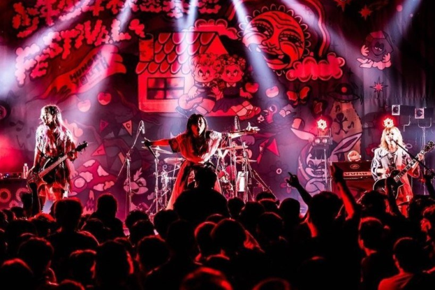 Ein Live-Auftritt der japanischen Rockband Korn auf der Bühne mit Musikern, die Gitarren spielen und ins Mikro singen, ein beleuchtetes Gemälde an der Wand dahinter und eine sichtbare Menge unten.