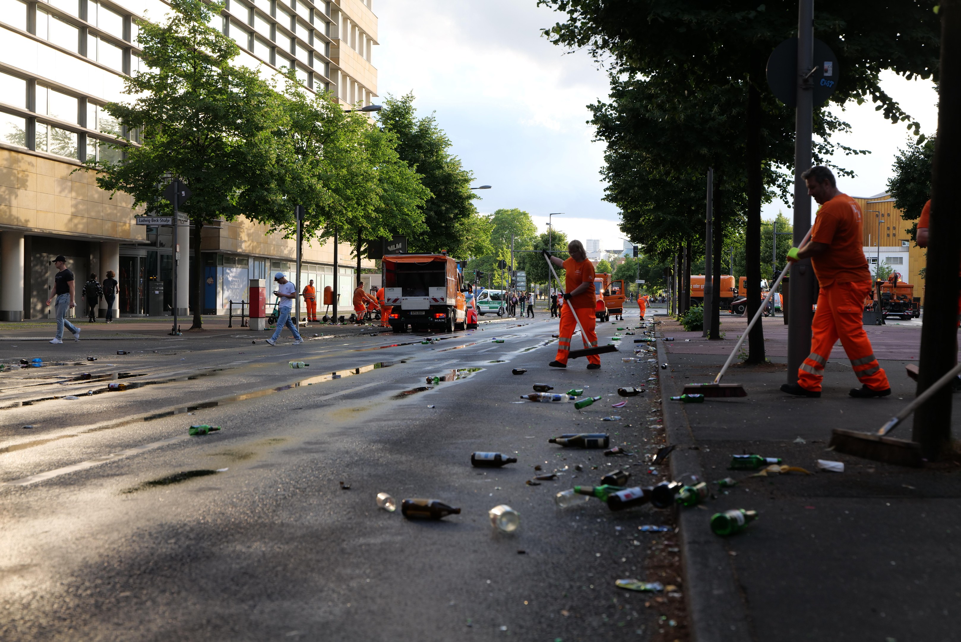 Eine Gruppe von Menschen in orangefarbenen Uniformen, die Müll von der Straßenseite entfernen, mit verstreutem Schutt, Bäumen, Polen und Fahrzeugen im Hintergrund.